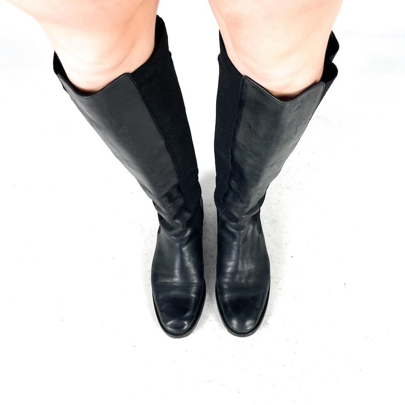 May include: A pair of knee-high black boots featuring a smooth leather front panel and a flexible, textured fabric back panel. The boots have a rounded toe, a flat sole, and extend up to the lower leg, showcasing a sleek, solid black design.