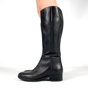 May include: A black leather knee-high boot with a rounded toe and a low heel. The boot has a smooth, polished finish and a simple, classic design. The shaft of the boot is tall and straight, and the sole is black.