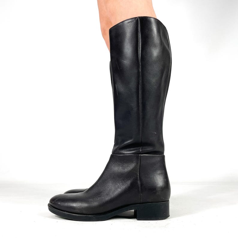 May include: A black leather knee-high boot with a rounded toe and a low heel. The boot has a smooth, polished finish and a simple, classic design. The shaft of the boot is tall and straight, and the sole is black.
