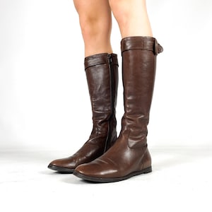 May include: Brown leather knee-high boots. One boot features a buckle detail at the top, the other a zip. The boots have a flat sole and a classic design. The boots are suitable for various outfits.