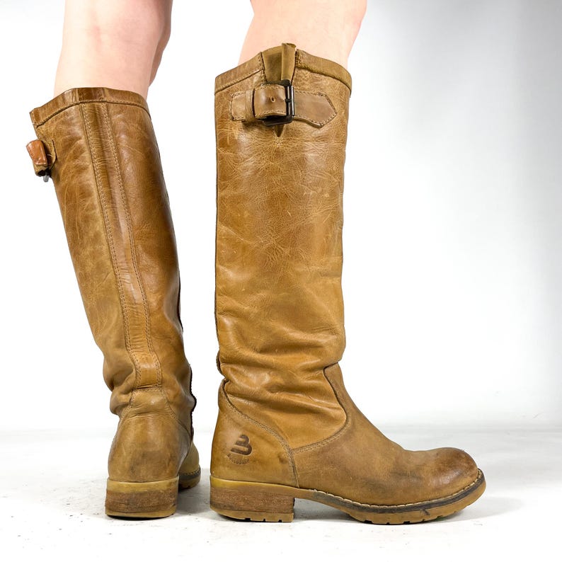 May include: A pair of tan leather knee-high boots. The boots have a worn, distressed appearance with buckles and a logo on the side. The soles are a light brown color.