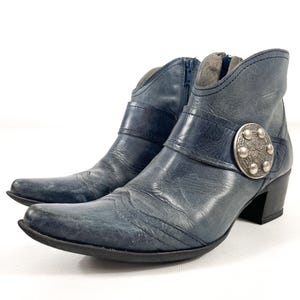 May include: Blue leather ankle boots with a pointed toe and a small heel. The boots have a decorative silver buckle and a zip closure. The leather has a distressed look. The boots are approximately 15 cm high.