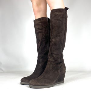 以下が含まれることがあります： ウェッジヒール付きの茶色のスエードニーハイブーツのペア。ブーツはジッパー開閉式で、丸いつま先です。スエード素材は柔らかく、質感のある外観です。ブーツは濃い茶色です。