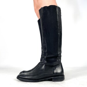 May include: A pair of black knee-high boots featuring a smooth leather toe and foot section, combined with a tall, flexible black fabric panel along the back of the shaft. The boots have a low, flat heel and a rounded toe design for a simple look.