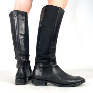 May include: A pair of black knee-high boots featuring smooth leather construction on the front and heel, complemented by a wide, flexible black elastic panel running along the back of the shaft. The boots have a low, flat heel and a rounded toe.