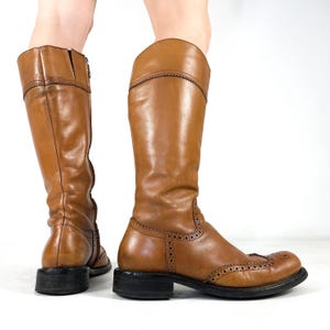 May include: A pair of knee-high tan leather boots featuring brogue detailing with decorative perforations along the toe cap and seams. The boots have a rounded toe, a low black stacked heel, and a vertical zip closure on the inner side.