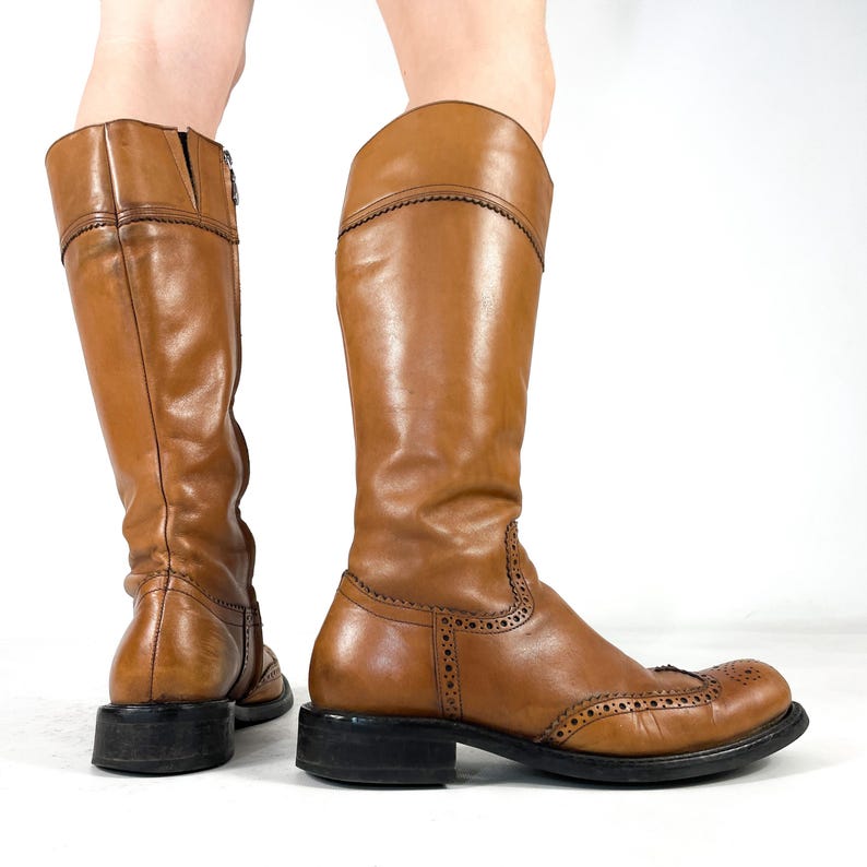 May include: A pair of knee-high tan leather boots featuring brogue detailing with decorative perforations along the toe cap and seams. The boots have a rounded toe, a low black stacked heel, and a vertical zip closure on the inner side.