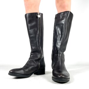 May include: A pair of black leather knee-high boots. The boots have a zipper closure on the side and a small heel. The boots are made of a smooth, shiny leather material. The boots are displayed on a white background.