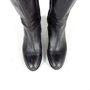 May include: A pair of black leather knee-high boots. The boots have a rounded toe and a slight heel. The leather appears worn, with some scuff marks visible. The boots are set against a white background.