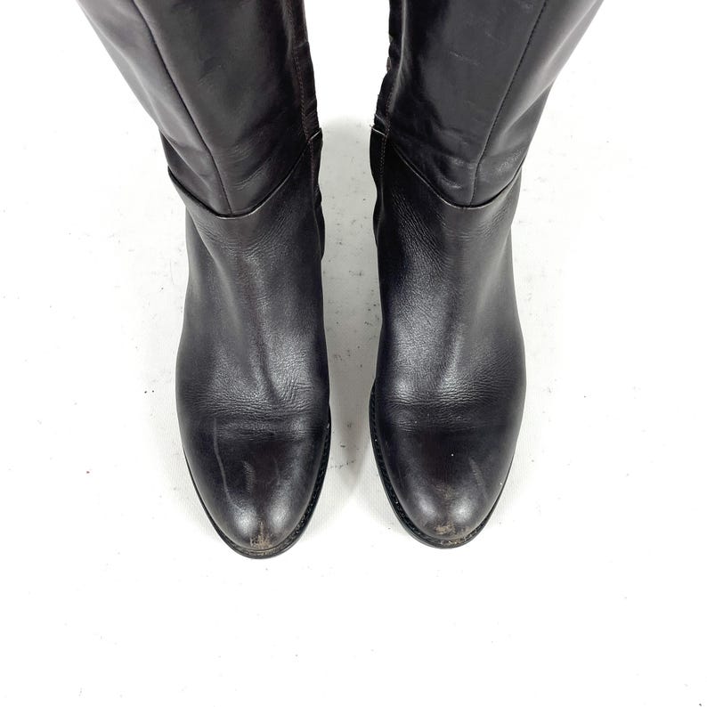 May include: A pair of black leather knee-high boots. The boots have a rounded toe and a slight heel. The leather appears worn, with some scuff marks visible. The boots are set against a white background.