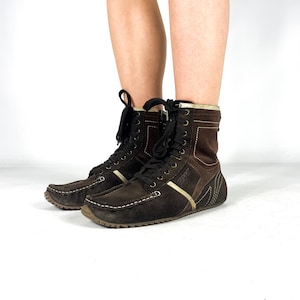 Pode incluir: Botas de tornozelo de camurça castanhas com atacadores pretos e acabamento creme. As botas têm biqueira estilo mocassim e sola de borracha. O nome da marca "Esprit" é visível na lateral da bota.