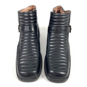 May include: A pair of black leather ankle boots. The boots have a square toe, a strap with a buckle, and a quilted design on the front. The boots are on a white background.