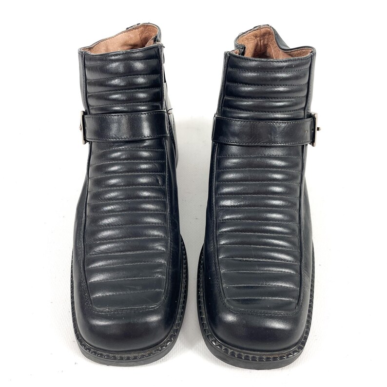 May include: A pair of black leather ankle boots. The boots have a square toe, a strap with a buckle, and a quilted design on the front. The boots are on a white background.