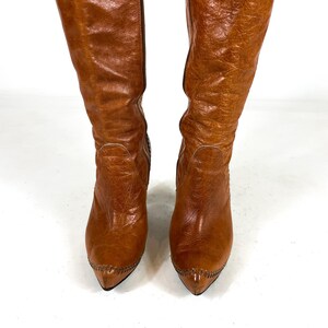 May include: A pair of knee-high, brown leather boots. The boots have a pointed toe and a textured surface. The boots are standing upright against a white background.