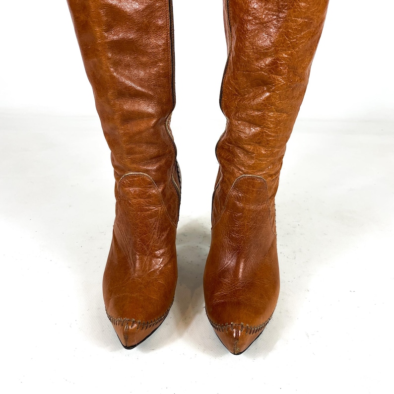May include: A pair of knee-high, brown leather boots. The boots have a pointed toe and a textured surface. The boots are standing upright against a white background.