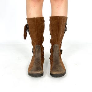 May include: Brown suede knee-high boots with buckle accents and a brown leather sole.