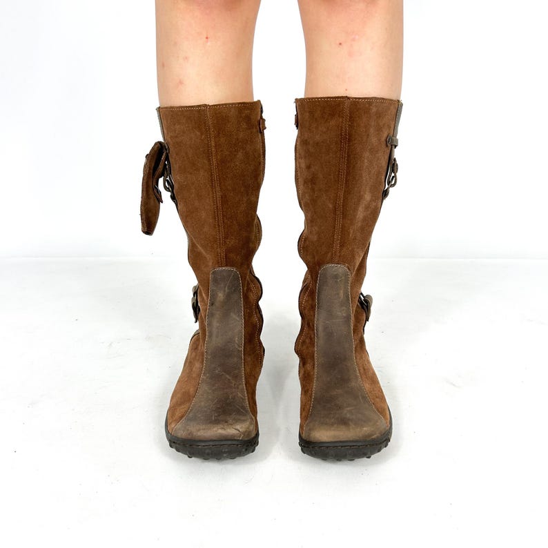 May include: Brown suede knee-high boots with buckle accents and a brown leather sole.