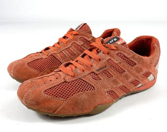 May include: Pair of orange suede and mesh sneakers with orange laces. The sneakers have the brand name "GEOX" on the side and a brown sole. The shoes are designed for athletic activities.