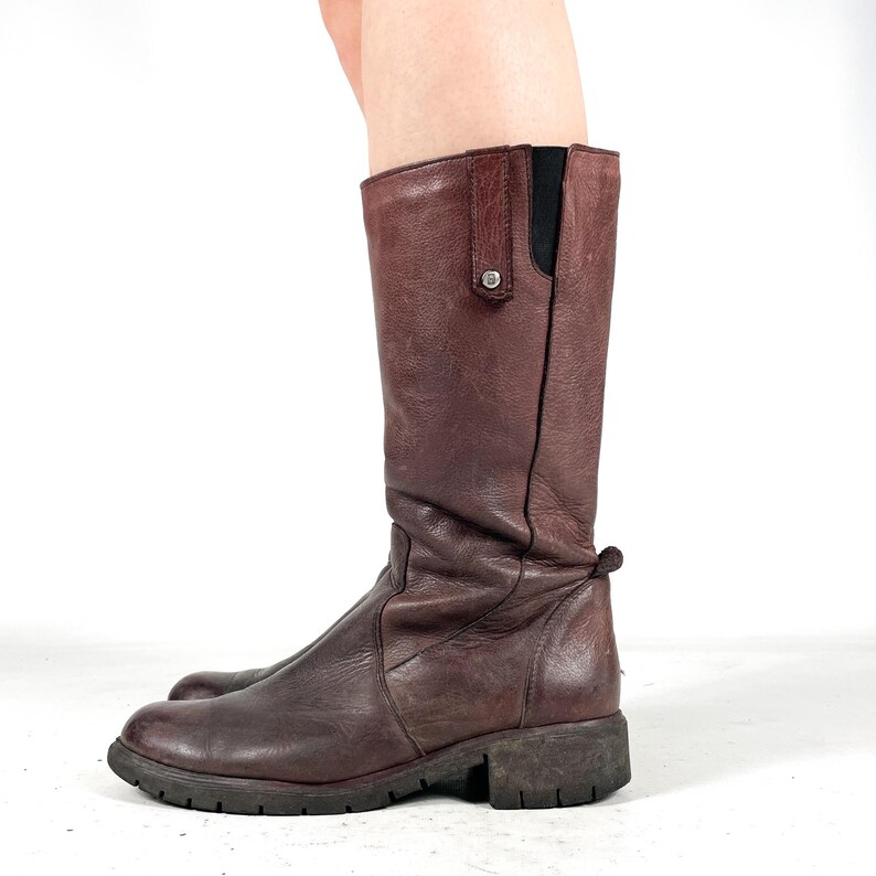 May include: A pair of brown leather knee-high boots. The boots have a rounded toe, a low heel, and a pull tab at the top. The leather has a distressed look.