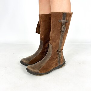 May include: Brown suede knee-high boots with a zipper on the inside of the leg and a buckle strap on the outside. The boots have a brown leather toe and heel.