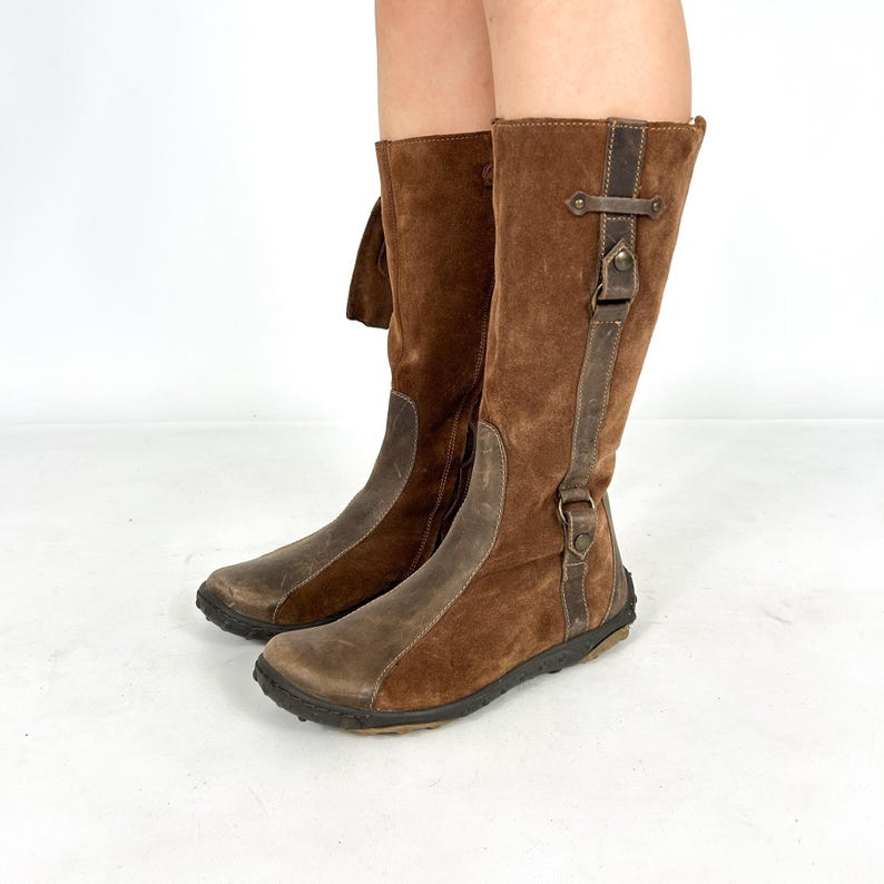 May include: Brown suede knee-high boots with a zipper on the inside of the leg and a buckle strap on the outside. The boots have a brown leather toe and heel.