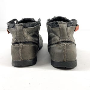 May include: A pair of gray leather ankle boots. The boots have a worn appearance with visible stitching and a black sole. The design includes a strap with an orange accent. The boots are viewed from the back.