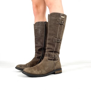 May include: Brown knee-high boots with buckle and strap details. The boots have a side zip and a flat sole. The boots are made of a textured material. The boots are worn by a person.