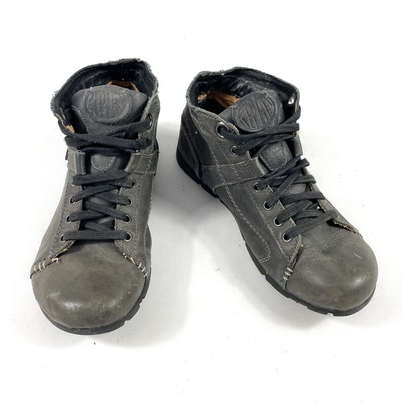 May include: A pair of gray leather high-top sneakers with black laces. The shoes have a worn appearance and feature stitching details and a logo on the tongue. The sneakers are designed for casual wear.