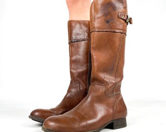 May include: A pair of brown leather knee-high boots. The boots have a worn appearance with visible creases and a buckle detail at the top. The boots are standing on a white surface.