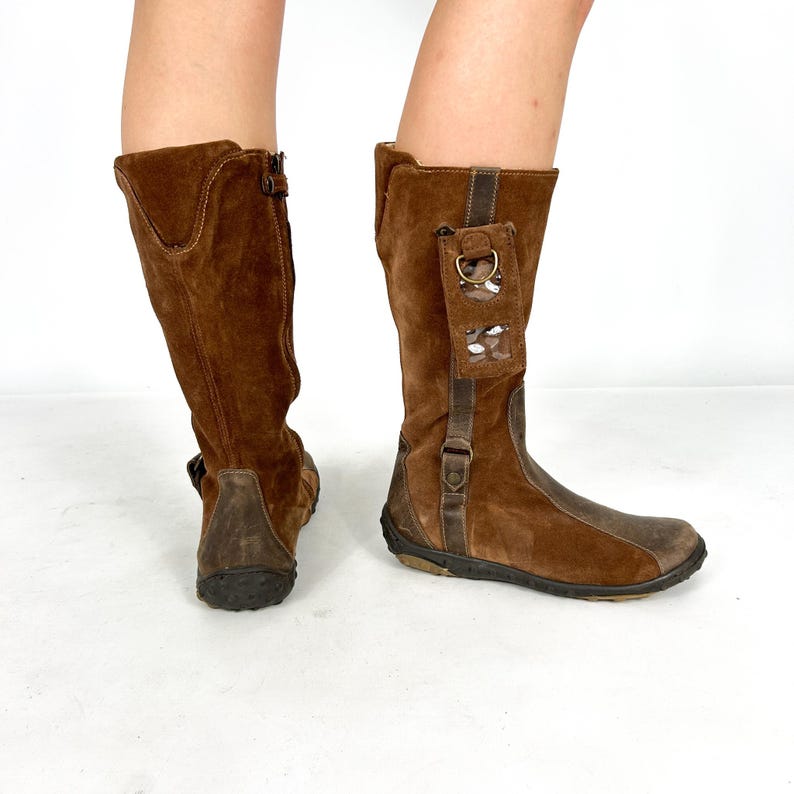 May include: Brown suede knee-high boots with a zipper on the side and a small pocket on the back of one boot.