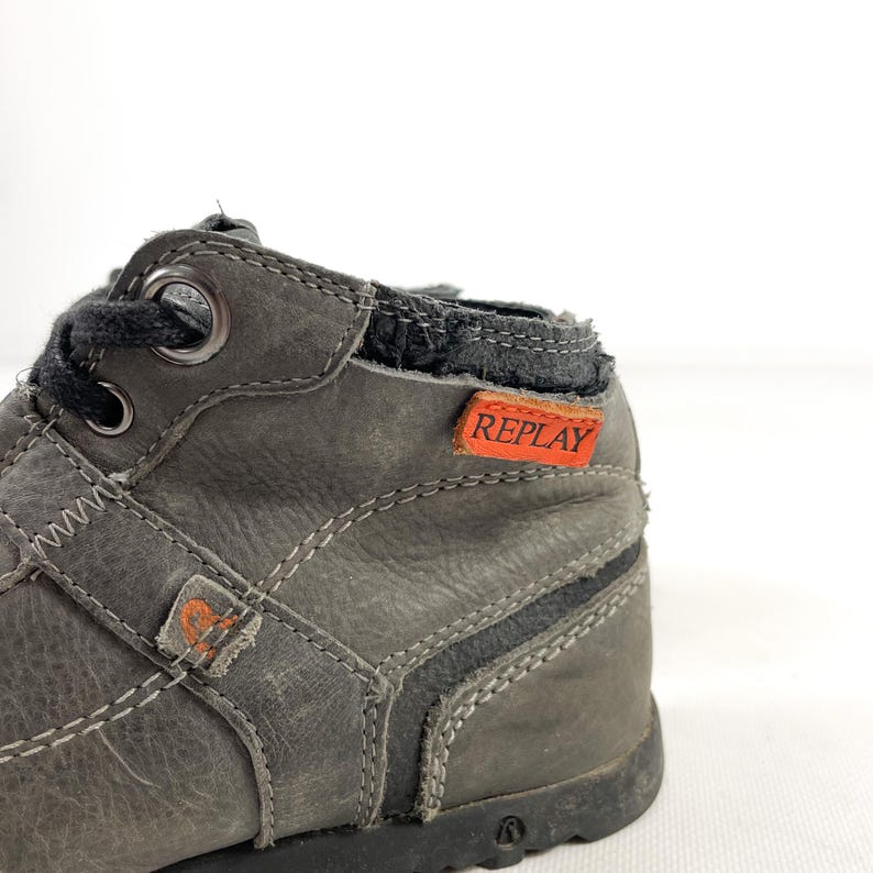 May include: Close-up of a gray leather boot with white stitching and black laces. The boot features an orange tag with the word "REPLAY" in white. The boot has a worn appearance.