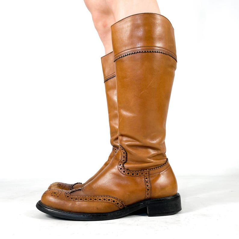 May include: A pair of knee-high tan leather boots featuring classic brogue detailing with decorative perforations along the toe cap and seams. The boots have a rounded toe, a low black heel, and a structured shaft that reaches the lower calf.