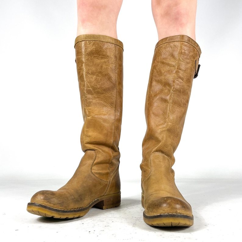 May include: A pair of knee-high, tan leather boots. The boots have a worn, distressed appearance with a buckle detail near the top. The soles are a contrasting light brown color. The boots are standing upright.