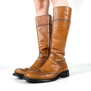 May include: A pair of knee-high tan leather boots featuring brogue detailing with perforated patterns along the toe cap and sides. The boots have a rounded toe, a low black heel, and a vertical zip closure on the inner side of each shaft.