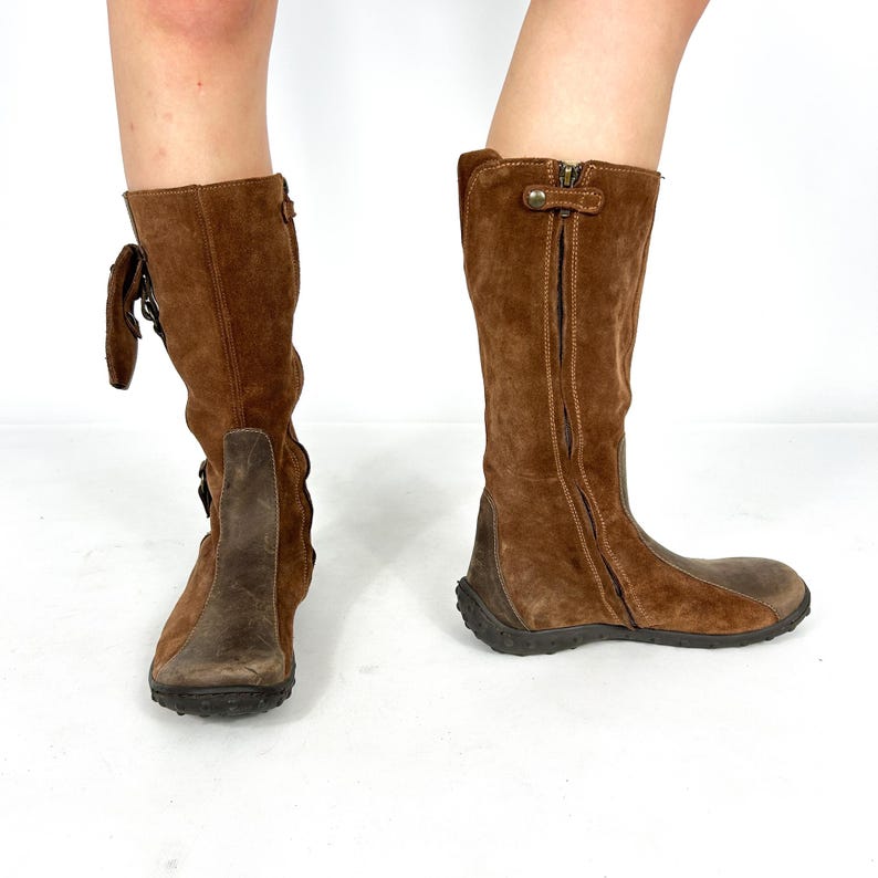 May include: Brown suede knee-high boots with a zipper on the side and a decorative buckle. The boots have a brown leather sole with a textured tread.