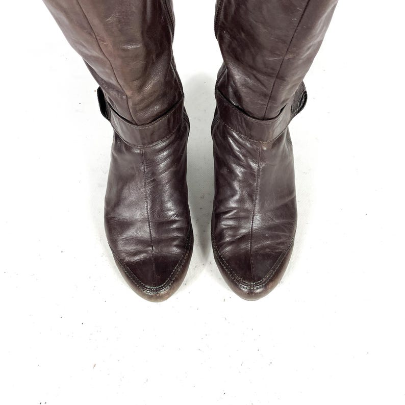 May include: A pair of knee-high brown leather boots. The boots have a pointed toe and a strap with a buckle around the ankle. The leather appears worn, with visible creases and variations in color.