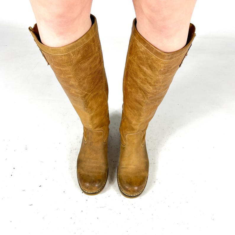May include: A pair of knee-high, tan leather boots. The boots have a distressed texture and a slight heel. The top of the boots have a decorative design. The boots are set against a white background.
