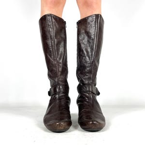 May include: A pair of knee-high brown leather boots. Each boot has a zipper on the side and a buckle strap near the ankle. The boots have a rounded toe and a slight heel.