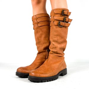 Könnte beinhalten: Ein Paar wadenhohe Stiefel aus hellbraunem Leder mit drei horizontalen Riemen und dunklen Metallschnallen an der Außenseite jedes Schafts. Die Stiefel haben eine abgerundete Zehenpartie und eine dicke, schwarze Profilsohle aus Gummi.