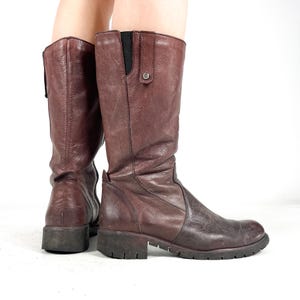 May include: A pair of knee-high, burgundy leather boots. The boots have a textured appearance, a small heel, and a side zipper. The boots are worn and show signs of use. The boots are on a white background.