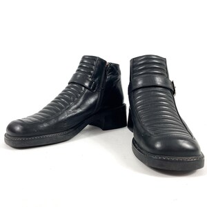 May include: A pair of black leather ankle boots with a ridged design. The boots have a side zipper, a buckle strap, and a thick sole. The boots are shown on a white background.