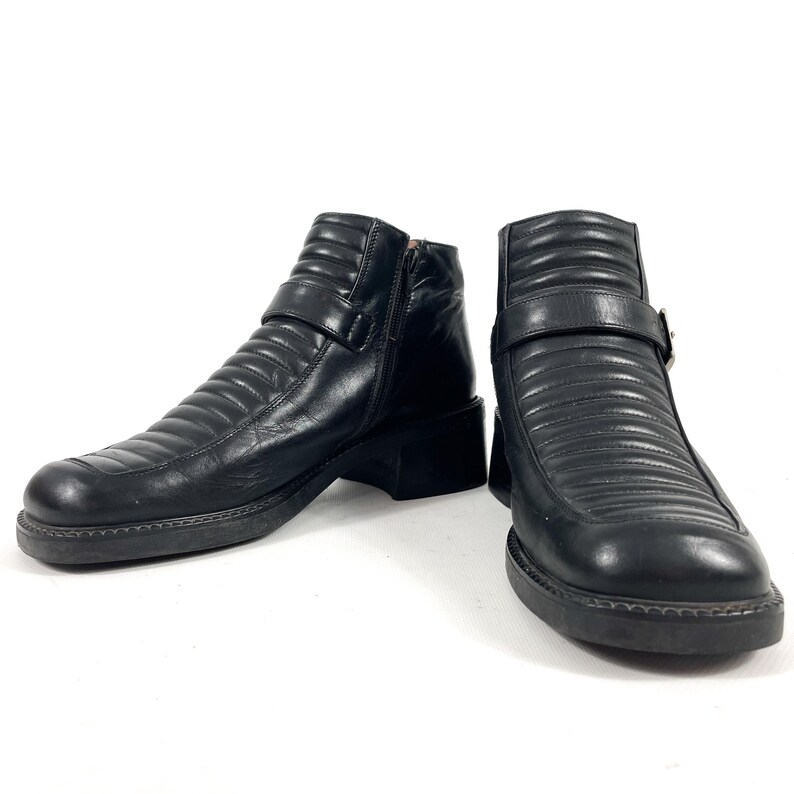 May include: A pair of black leather ankle boots with a ridged design. The boots have a side zipper, a buckle strap, and a thick sole. The boots are shown on a white background.