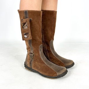 May include: Brown suede knee-high boots with a zipper on the inside of each boot. One boot has a clear plastic pocket with a metal ring on the side.