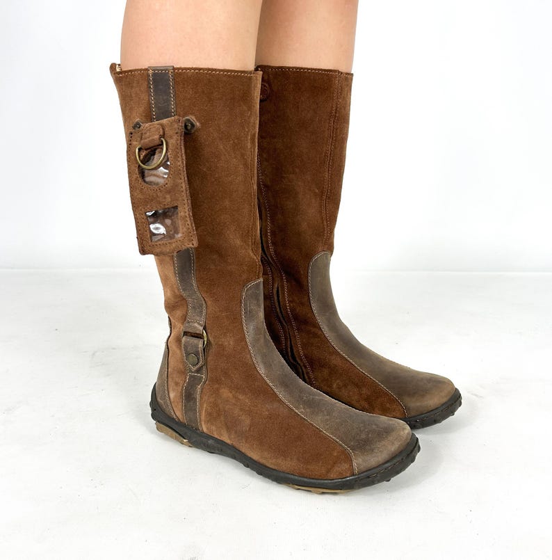 May include: Brown suede knee-high boots with a zipper on the inside of each boot. One boot has a clear plastic pocket with a metal ring on the side.