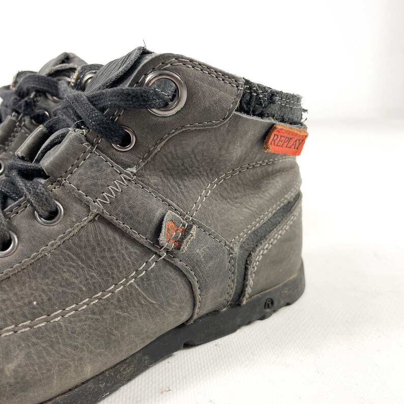 May include: Close-up of a pair of gray leather high-top sneakers. The sneakers have black laces, metal eyelets, and white stitching. A red tag with the word "REPLAY" is visible on the side. The shoes have a worn, distressed appearance.