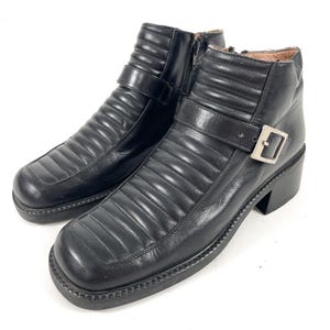 May include: A pair of black leather ankle boots with a square toe and a block heel. The boots feature a quilted design, a side zipper, and a buckle strap. The boots have a lug sole.