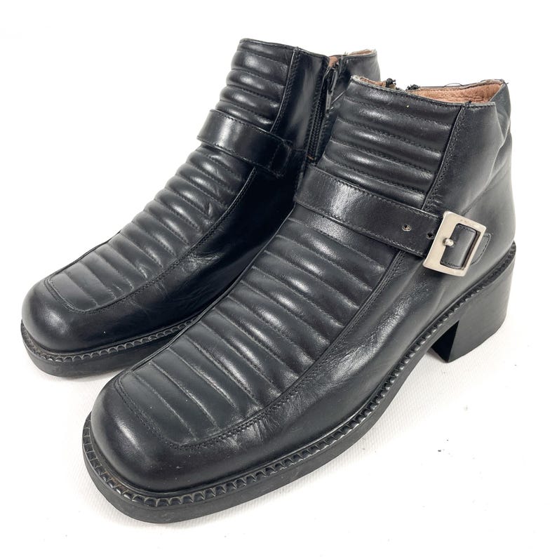 May include: A pair of black leather ankle boots with a square toe and a block heel. The boots feature a quilted design, a side zipper, and a buckle strap. The boots have a lug sole.