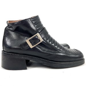 May include: Black leather ankle boots with a square toe and a low block heel. The boots feature a silver-tone buckle closure and quilted detailing on the upper. The boots are a classic style, suitable for various outfits.