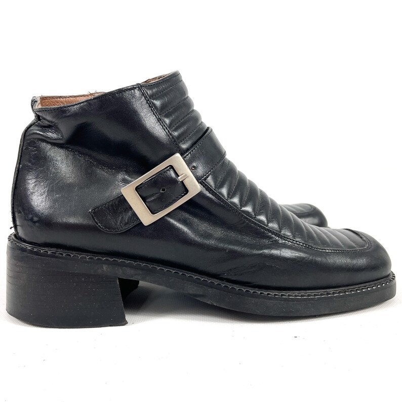 May include: Black leather ankle boots with a square toe and a low block heel. The boots feature a silver-tone buckle closure and quilted detailing on the upper. The boots are a classic style, suitable for various outfits.