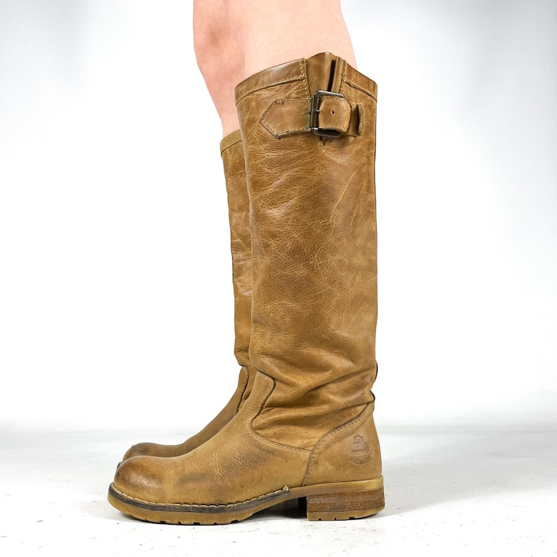 May include: A pair of tan leather knee-high boots. The boots have a distressed appearance, with a buckle detail at the top and a rounded toe. The soles are a lighter brown, and the boots appear worn.